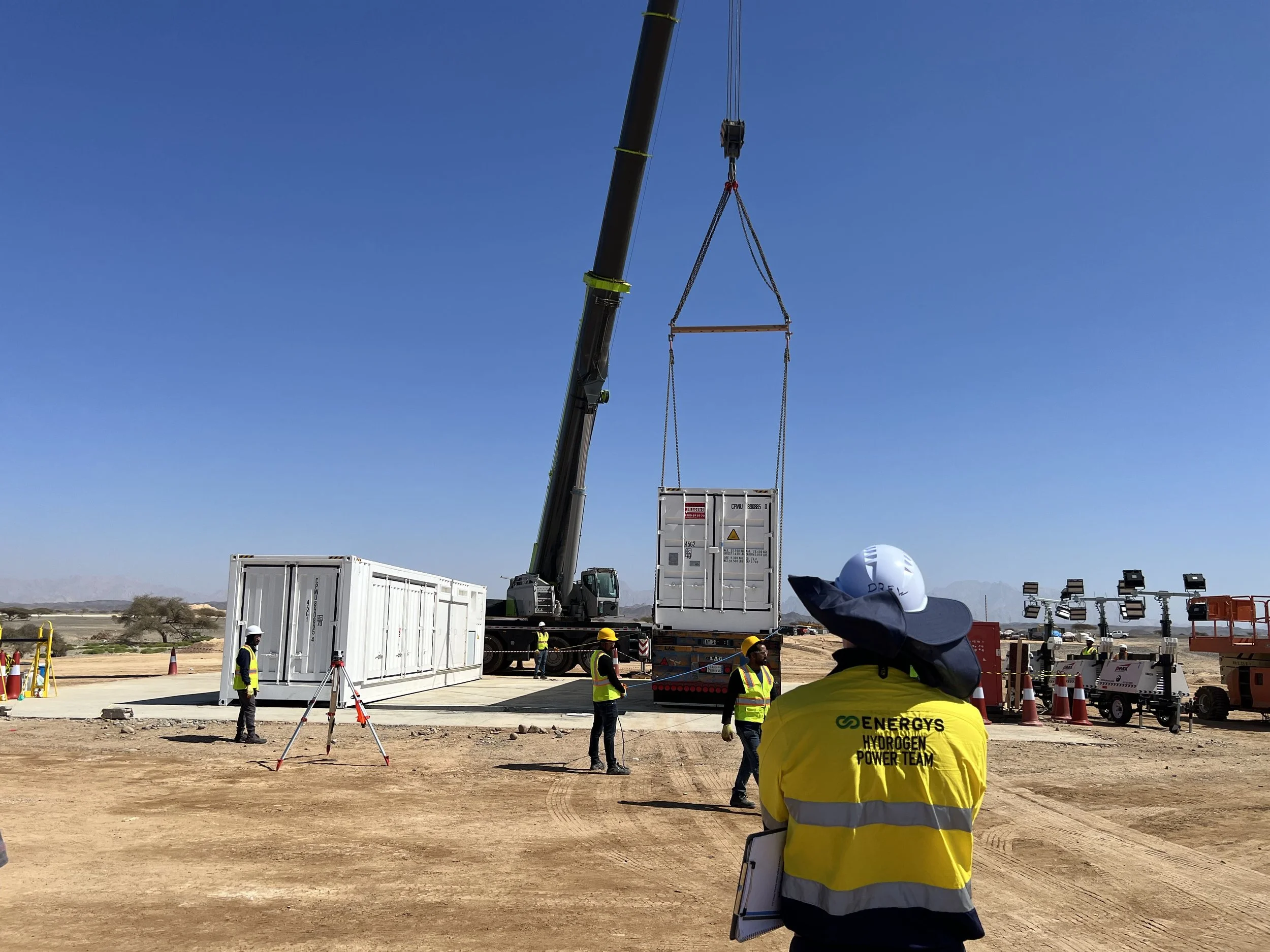Construction workers in safety vests and helmets working on a hydrogen power project in a desert landscape. A crane is lifting a container labeled 'Hydrogen' and 'Energys.' The scene includes various construction equipment and vehicles.
