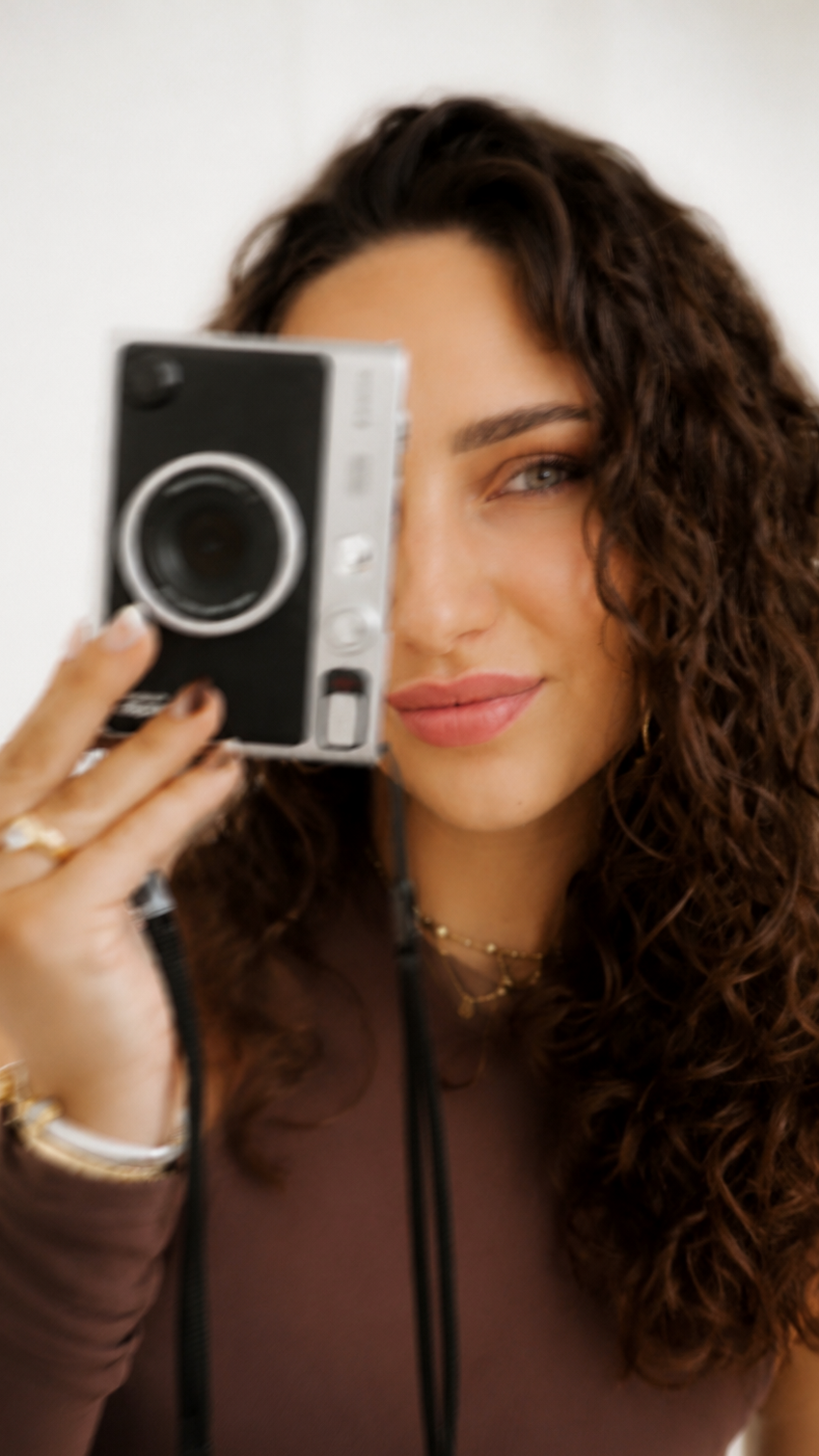 Vrouw met bruin haar en make-up houdt een vintage camera dicht bij haar gezicht, lachend en naar de camera kijkend.