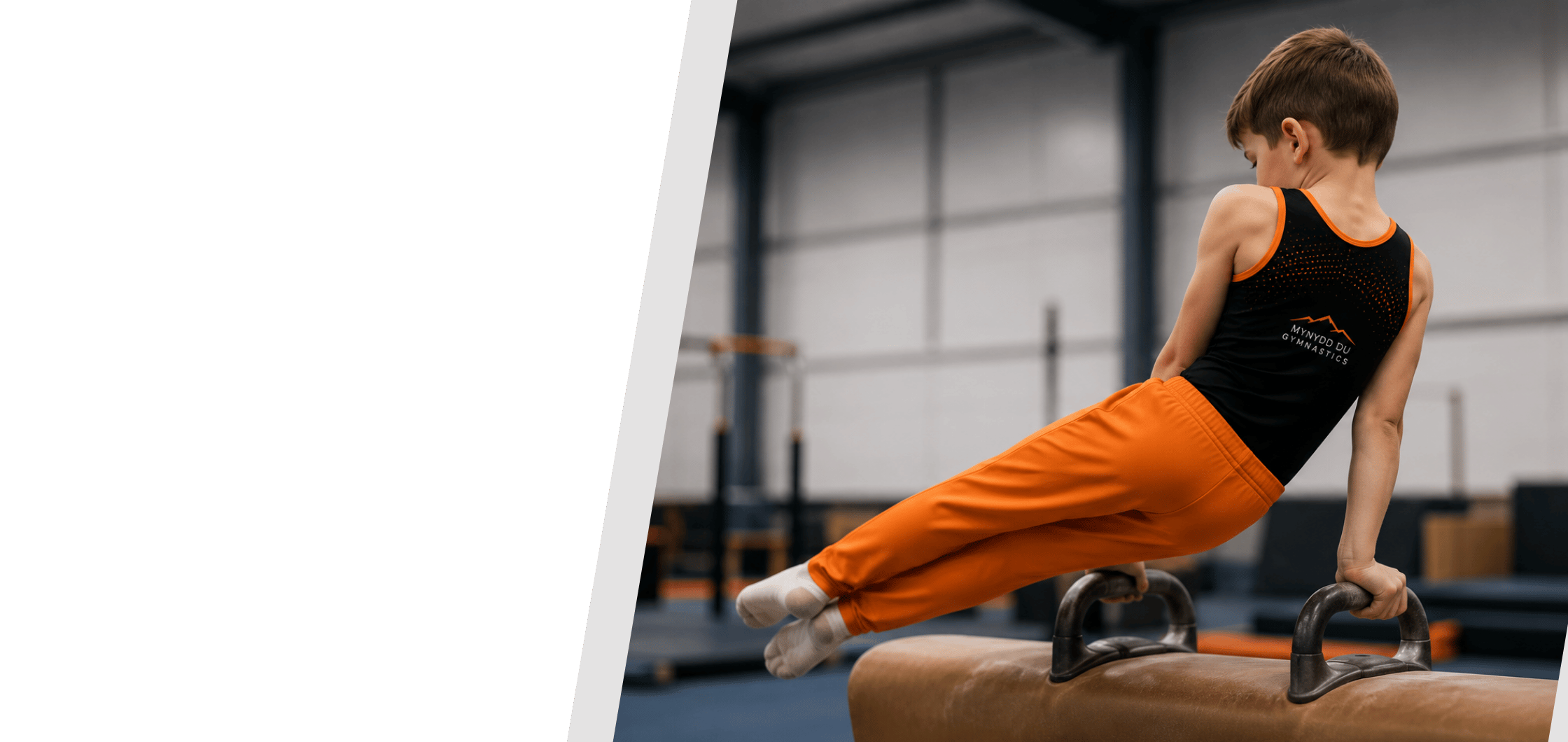 A young male gymnast in a black and orange uniform performs an exercise on parallel bars in an indoor gym.