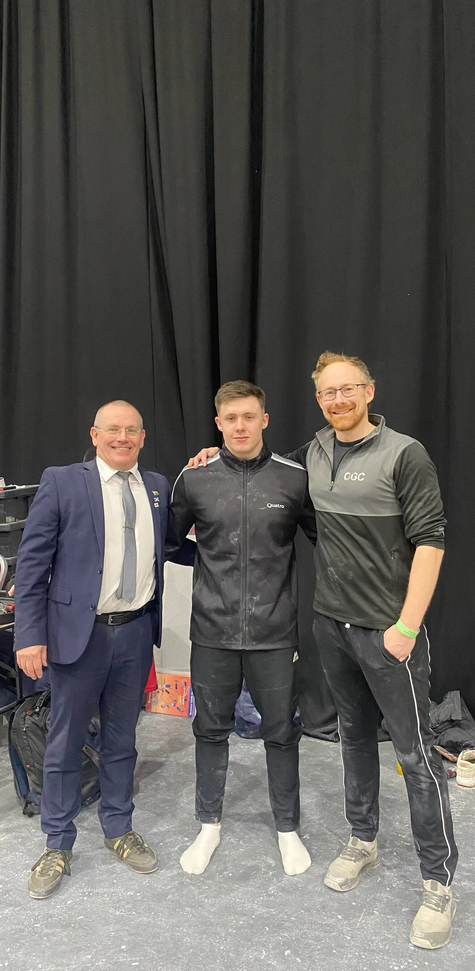 Three men standing together, smiling, in front of a black curtain. The man in the middle is wearing a black athletic jacket and black pants with white socks, and appears to be a cyclist. The man on the left is dressed in a navy suit with a large tie,