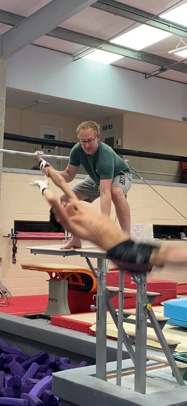 A man and a dog practicing in a dog training or agility gym; the dog is mid-air, jumping off a table, and the man is crouched on the table, holding the dog with gloves.