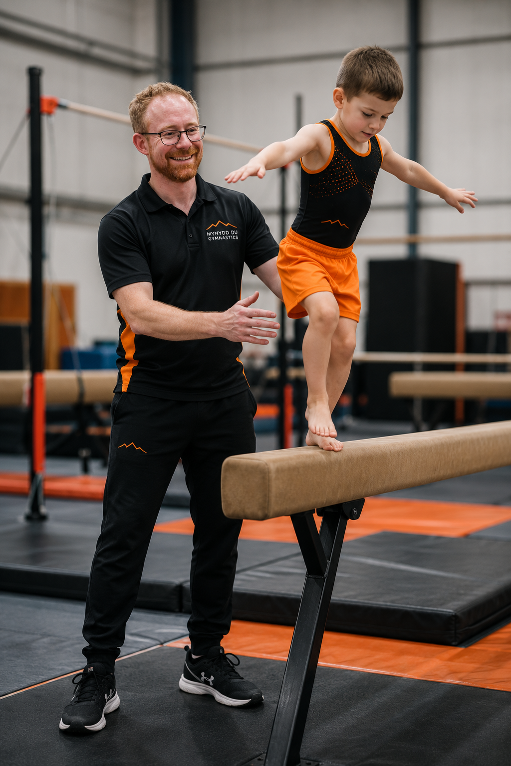 A young boy practicing gymnastics on a balance beam with a coach guiding him in an indoor gym. The coach is smiling and wearing a black shirt with orange accents, and the boy is wearing an orange and black athletic outfit.