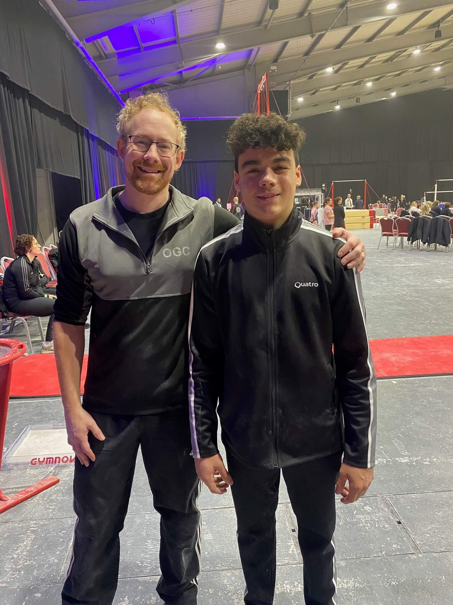 Two men standing together inside a gym, both wearing black and gray jackets, with one man having curly hair and the other with glasses and a beard. They are smiling and appear to be happy.