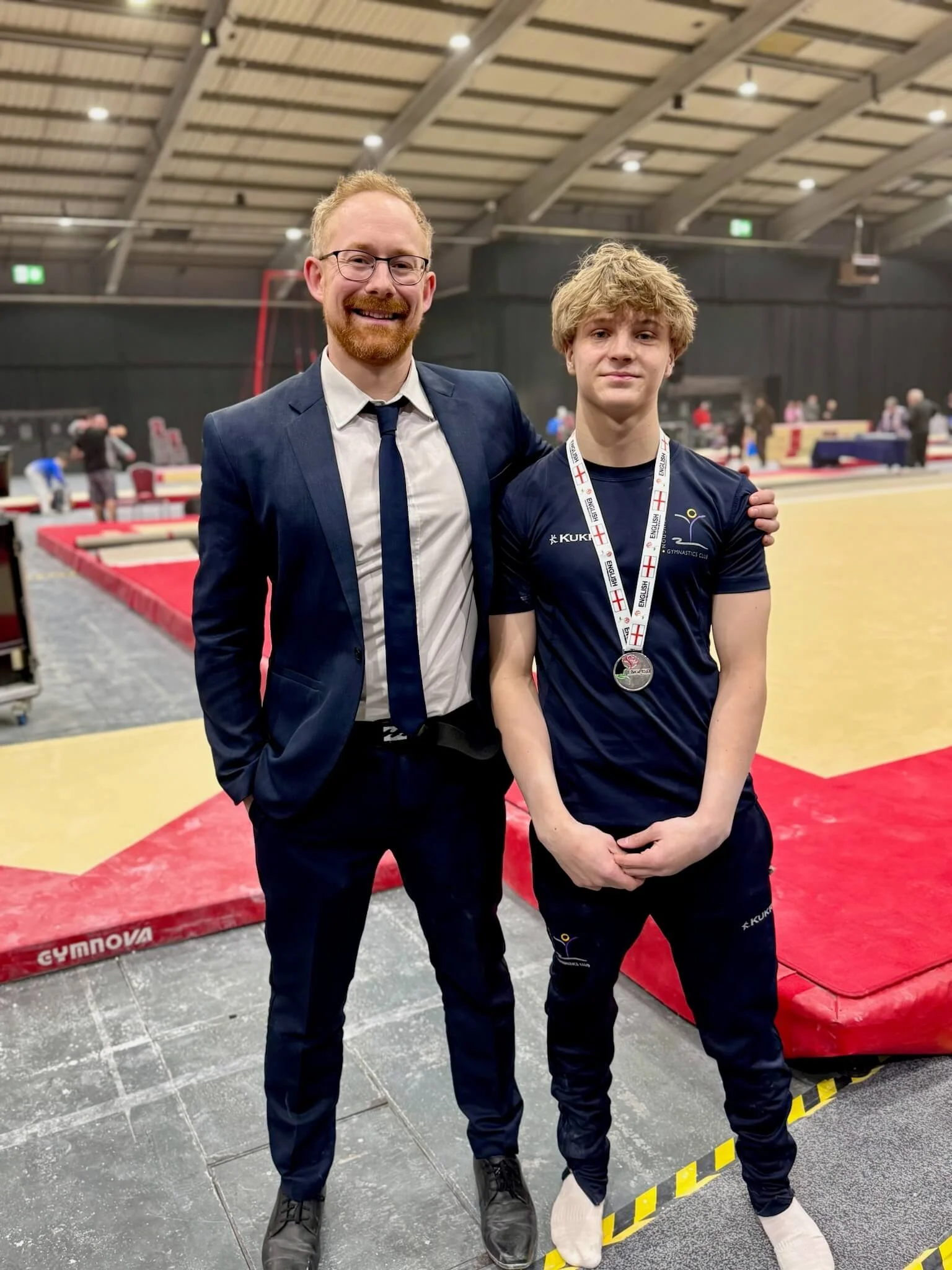 A man in a dark suit and glasses standing next to a young athlete wearing a sports uniform and a medal at an indoor gymnastics venue.