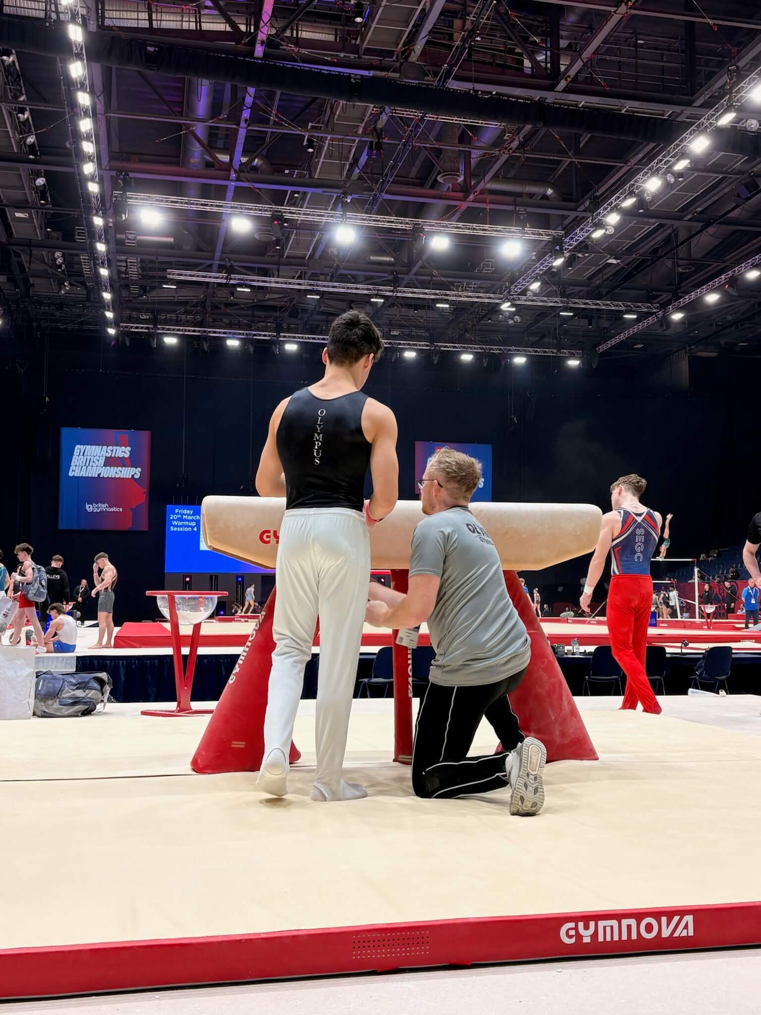 A gymnast practicing on a vaulting table at a gymnastics competition, with a coach kneeling beside him and other gymnasts in the background. Signs indicate it is the British Gymnastics Championships.