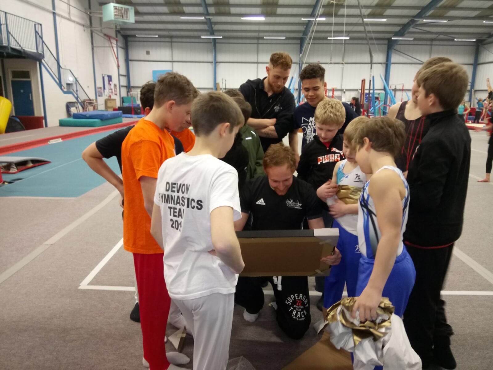 A group of young gymnasts and their coaches gathered around a table in an indoor gymnasium, looking at a clipboard or a tablet, likely reviewing scores or results.