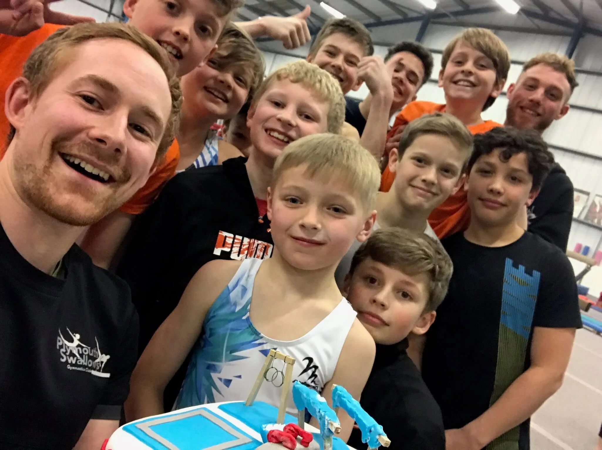 A group of children and two adults smiling and celebrating at an indoor event, with a birthday cake or a decorated dessert in the foreground.