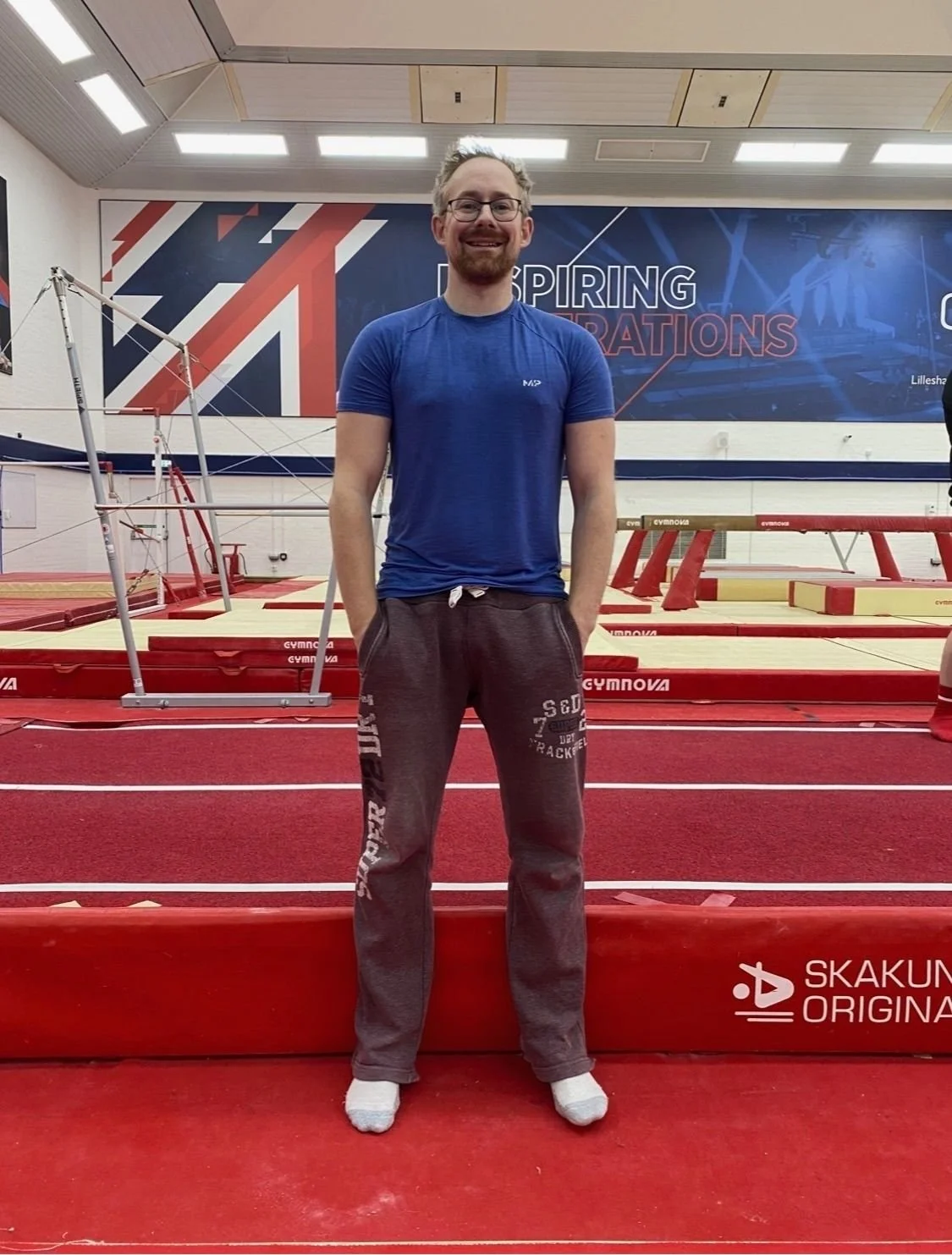 A smiling man with glasses and a beard standing on a red gymnastics mat inside a training facility. He is wearing a blue athletic shirt and gray sweatpants with text, with hands in his pockets. The background shows gymnastics equipment like parallel bars, a springboard, and a large banner with geometric designs and the words "Spring Celebrations."