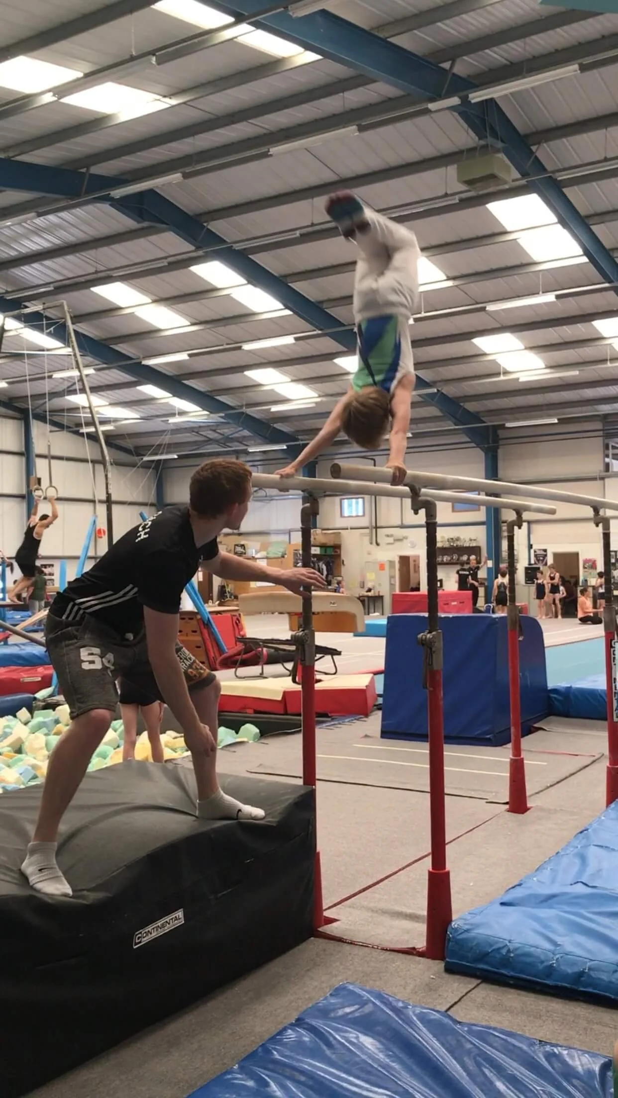 A young gymnast performing a handstand on parallel bars in a gymnastics training facility, watched by a coach.