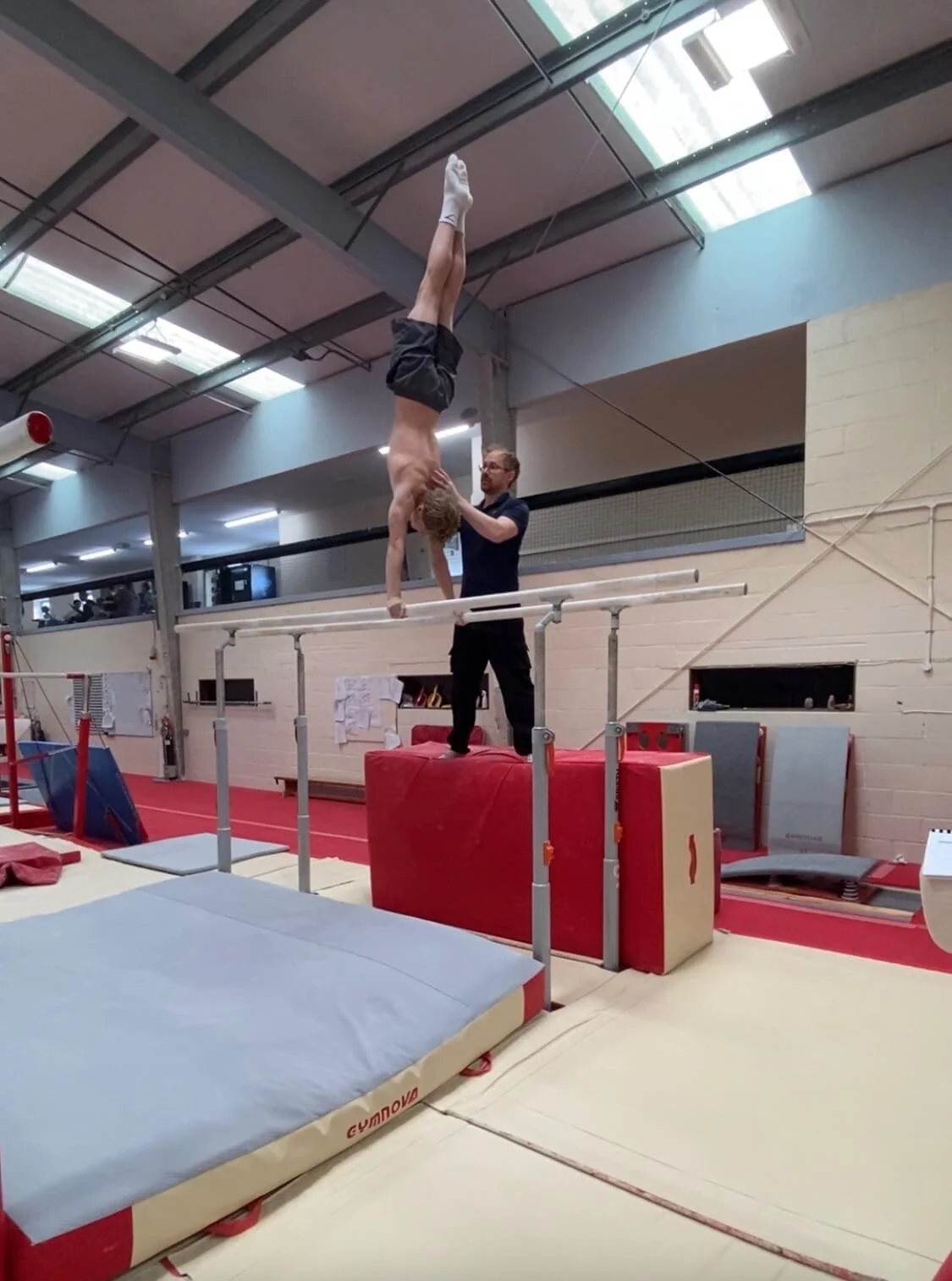 A gymnast is performing an upside-down handstand on the parallel bars, with a coach supporting him from below, in a gymnastics training facility.