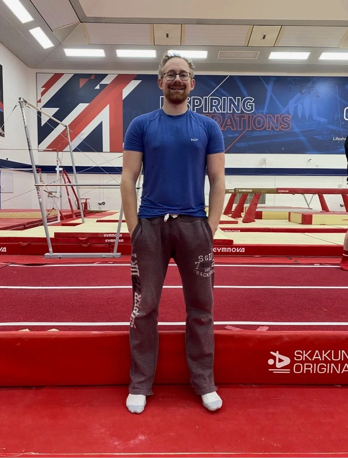 A man standing on a red gymnastics floor in a gymnasium, smiling at the camera. He is wearing a blue t-shirt, gray sweatpants, and white socks. Gymnastics equipment, including a balance beam and parallel bars, are visible in the background, with a la