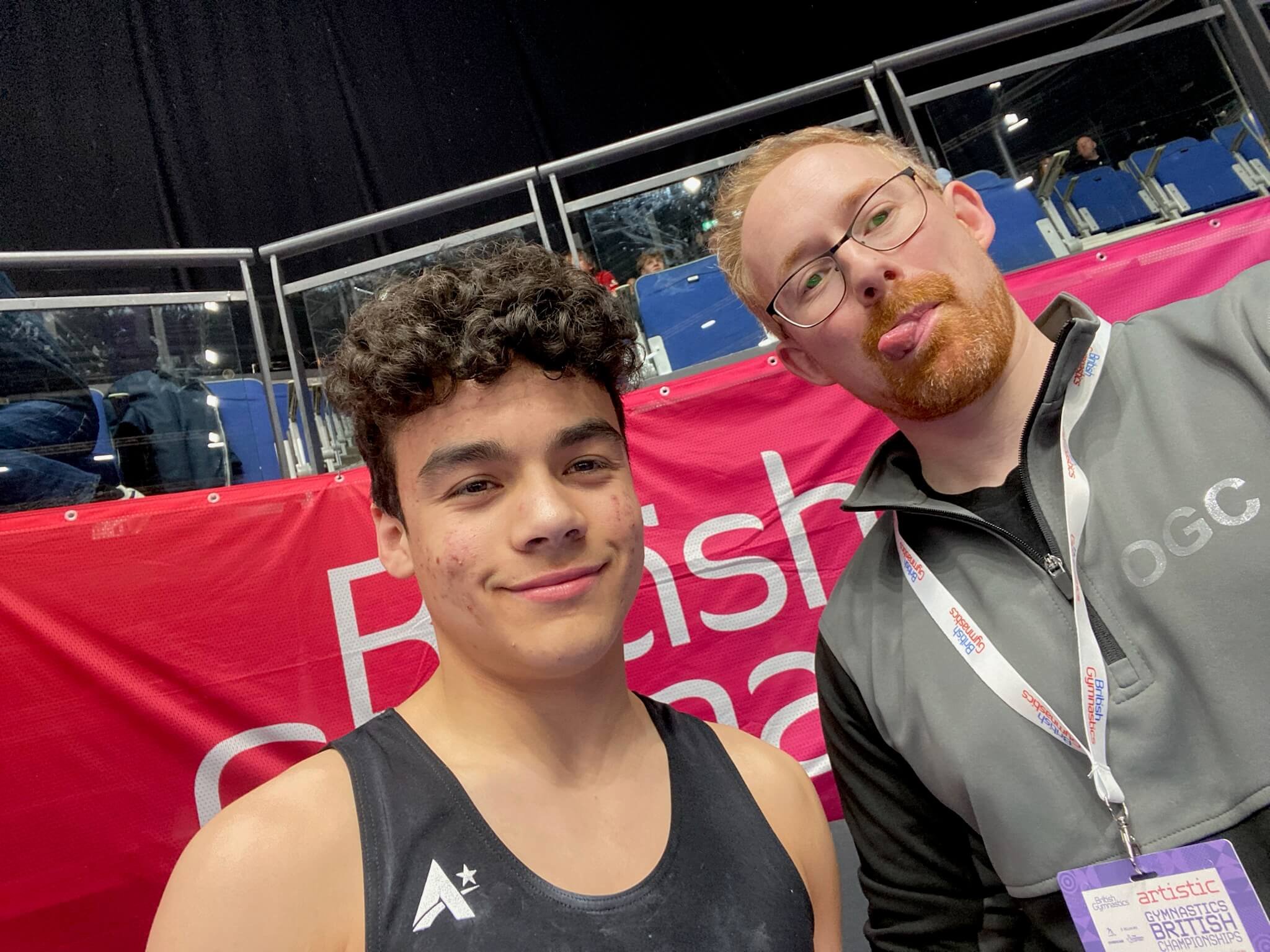 Two young men taking a selfie at a sports event. The man on the right has glasses and red hair and beard, and is sticking out his tongue. The man on the left has curly dark hair and is smiling. They are seated in an arena with blue seats and are in f