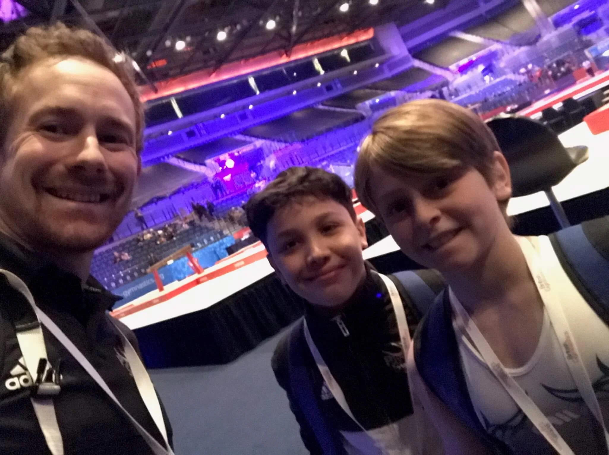 A close-up photo of three young boys smiling at an indoor event, with a large, colorful arena or stadium structure illuminated in red, purple, and blue lights in the background.