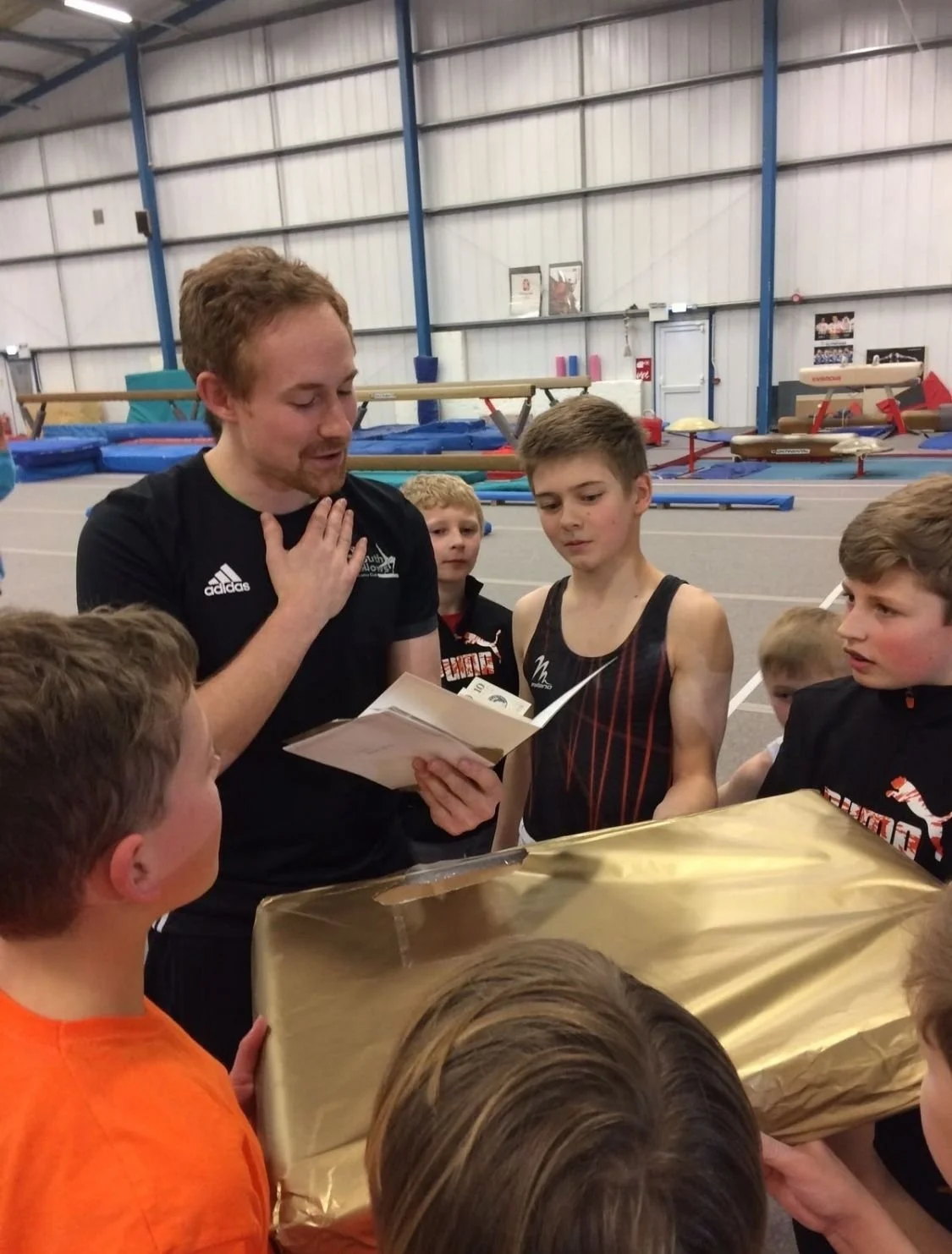 A coach gives a speech to young athletes, reading from a paper, inside an indoor gymnastics facility. The athletes are gathered around a table with a gold wrapped present, some eyeing the coach. The background features gymnastics mats and equipment.