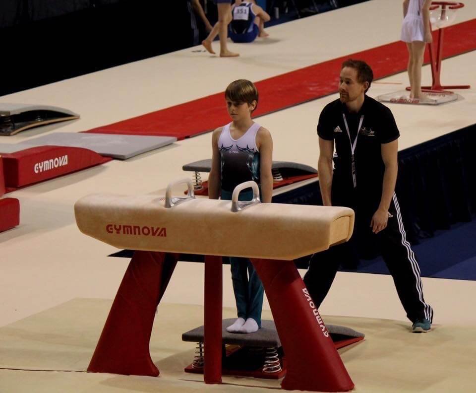 A young female gymnast preparing for her routine on the vault apparatus, with a coach standing nearby at an indoor gymnastics competition.