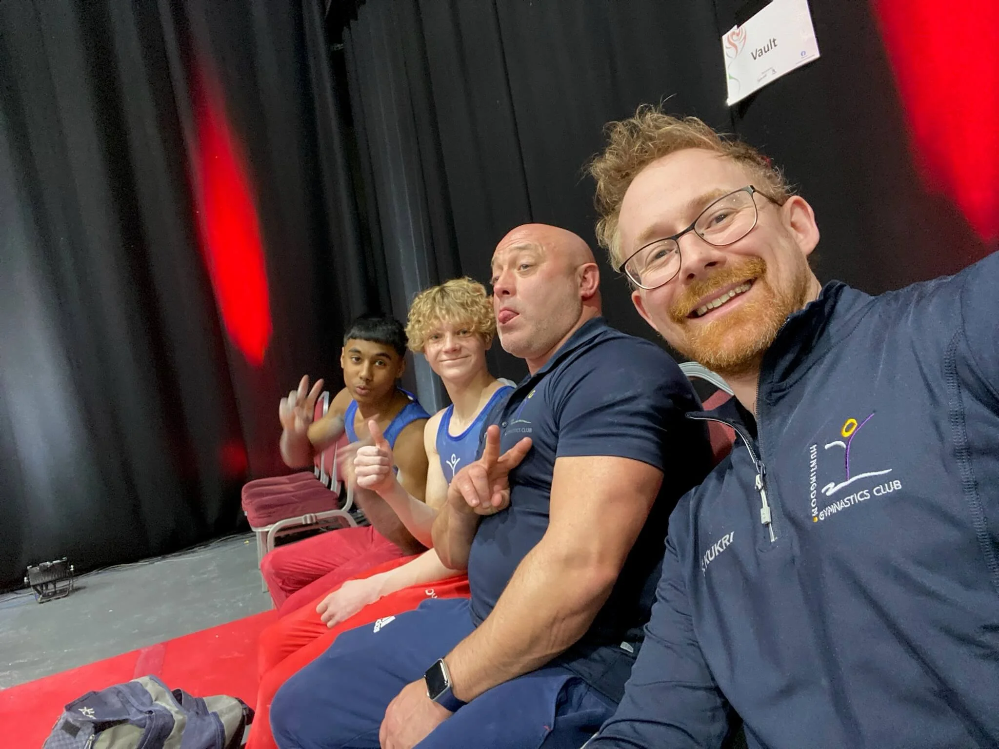 A group of five people seated in a row on a red bench at an indoor sports arena, with black curtains behind them and a sign that reads 'Vault' above. The group includes four men and one woman, all smiling or making playful gestures towards the camera