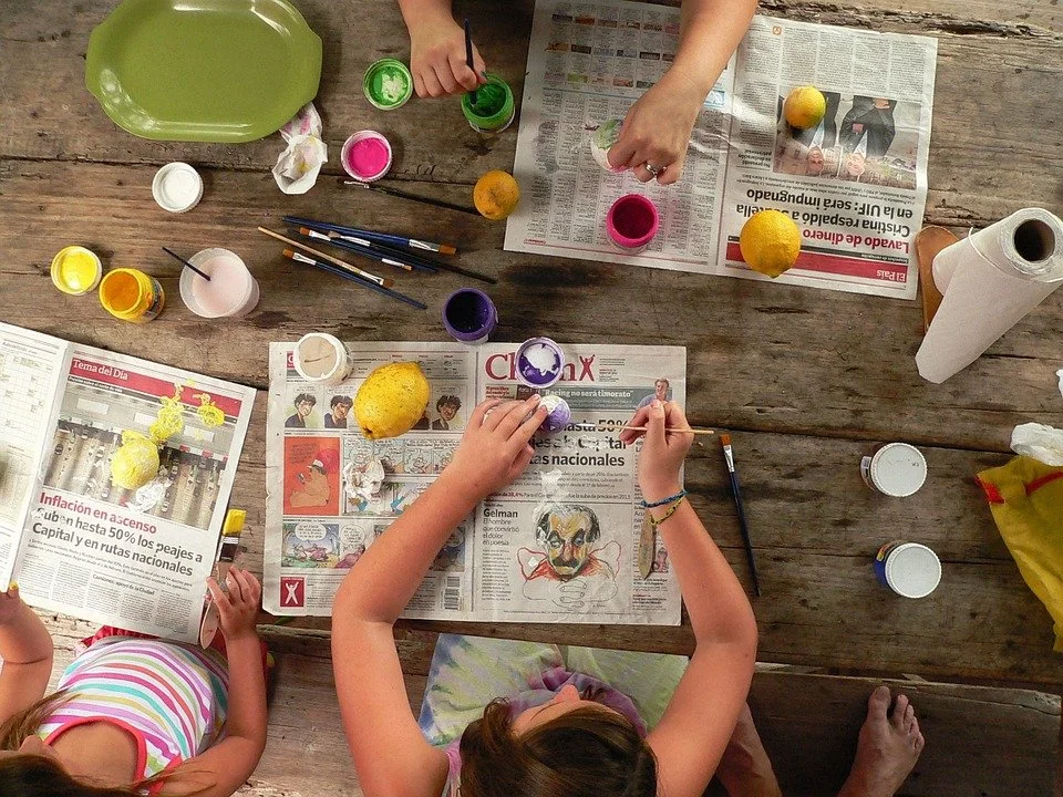 Several children are painting and drawing on Sunday newspapers spread on a wooden table. They have jars of paint, brushes, and lemons used as art tools. The scene shows a creative art activity.