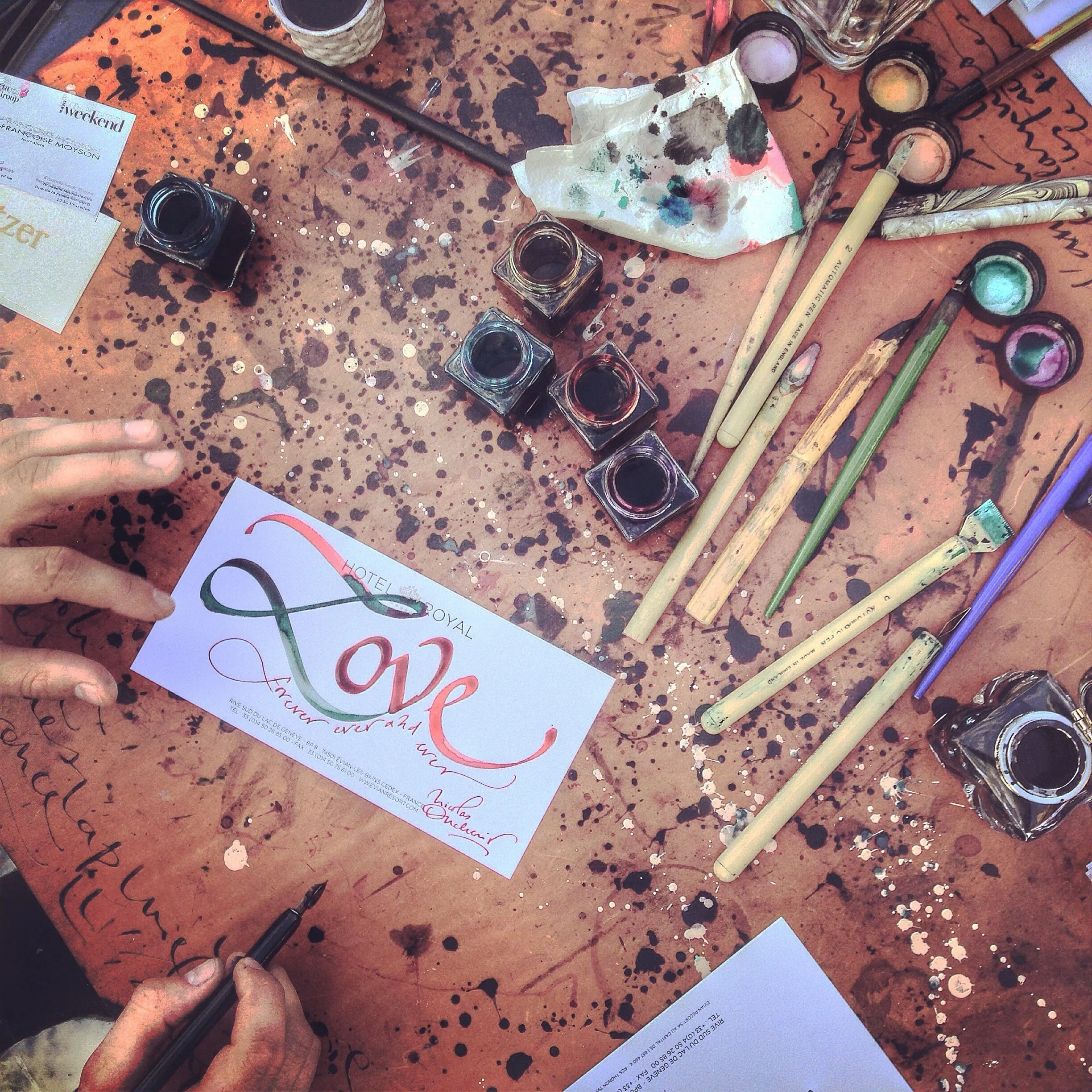 A tabletop covered with spilled ink, paint, and confetti. Several bottles of ink and paintbrushes are scattered across the table, along with a crumpled piece of paper with watercolor stains. There is a partially visible flyer or card with artistic writing and colorful script, and a person's hand holding a black pen in the lower left corner.