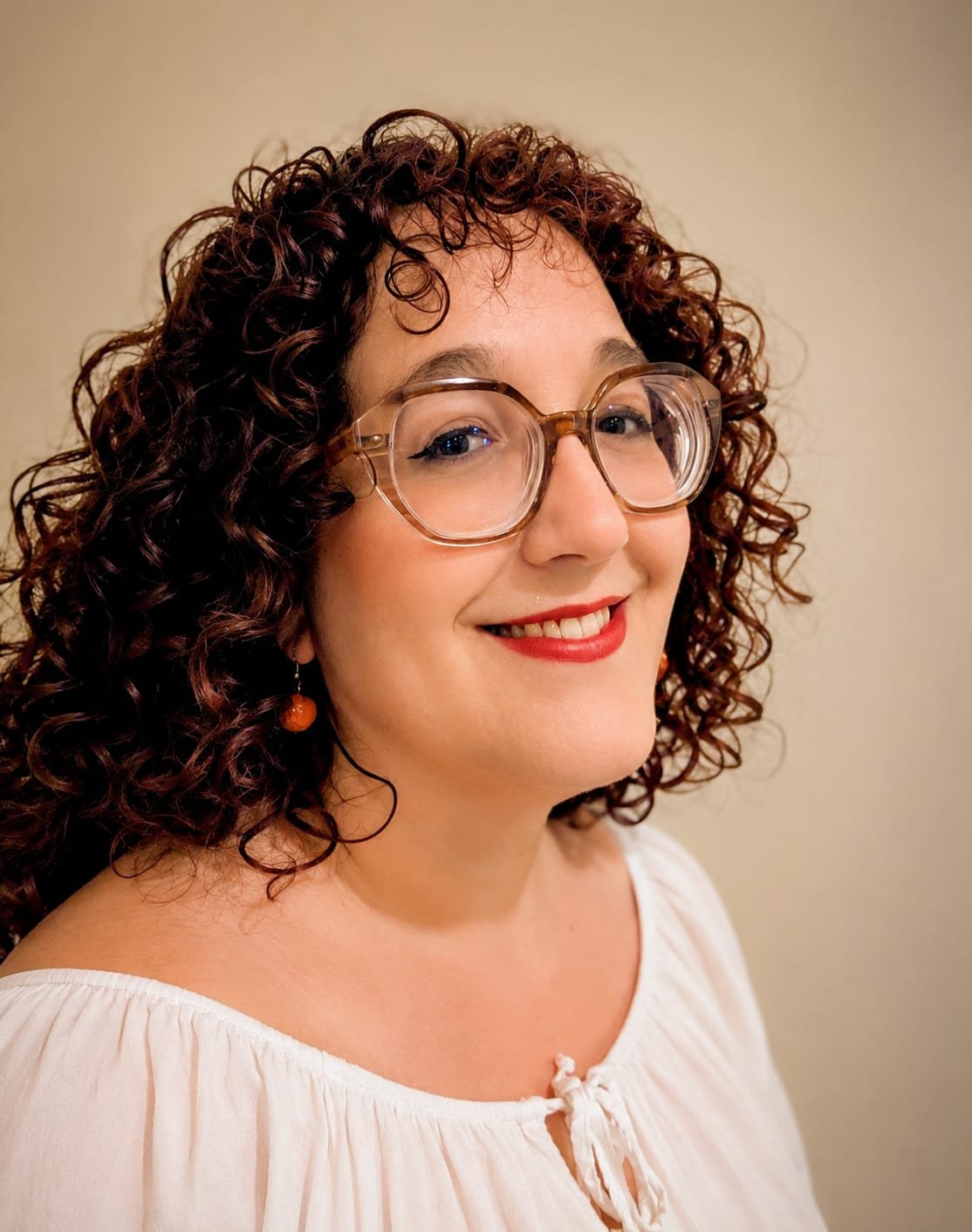 A woman with curly dark brown hair, wearing large clear glasses, red lipstick, a white blouse, and orange earrings, smiling against a beige background.