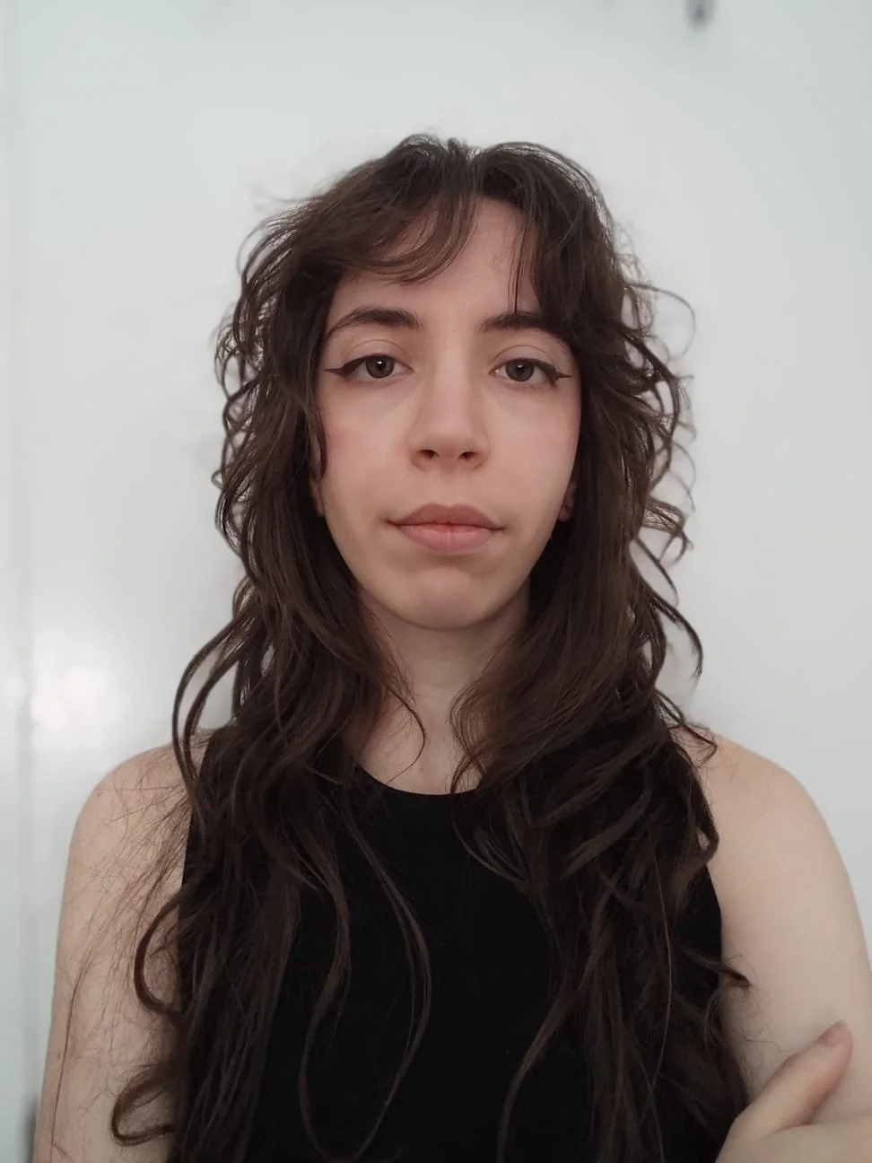 A young woman with wavy brown hair and eyeliner wearing a black sleeveless top standing in front of a plain white wall.