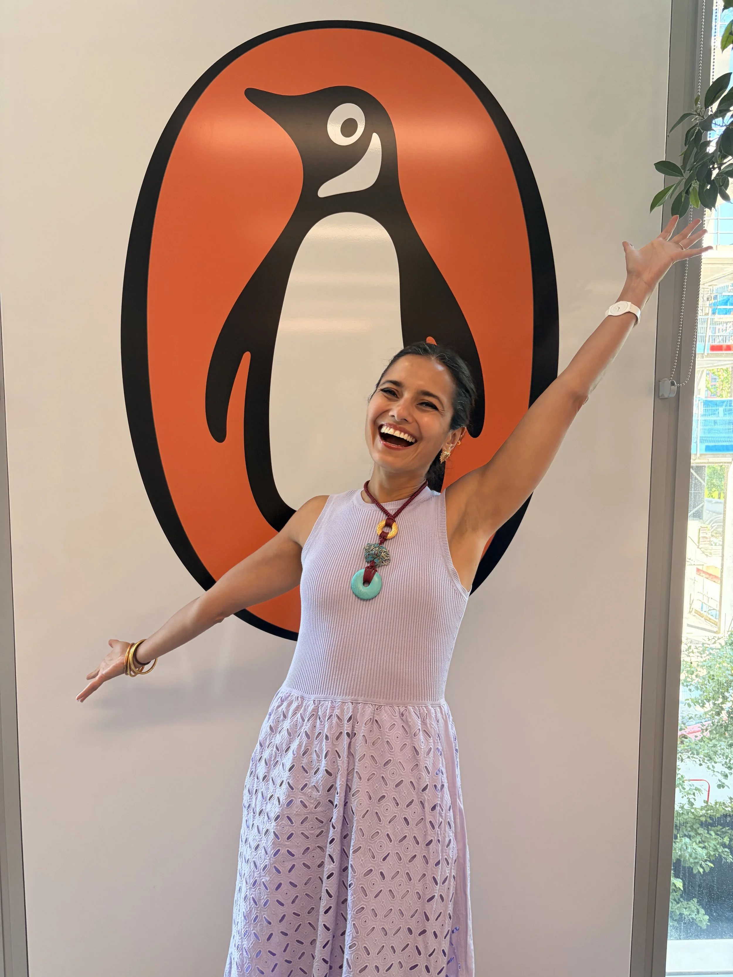 A woman wearing a sleeveless white top and pink patterned pants, smiling and raising one arm, standing in front of a large orange and black penguin illustration on the wall.