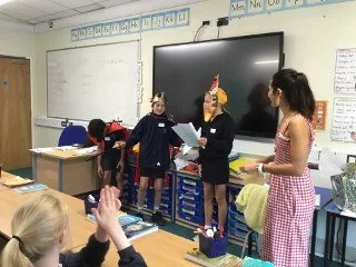 Children in a classroom wearing unicorn and unicorn headbands, with one girl in a checkered dress, engaged in a classroom activity.