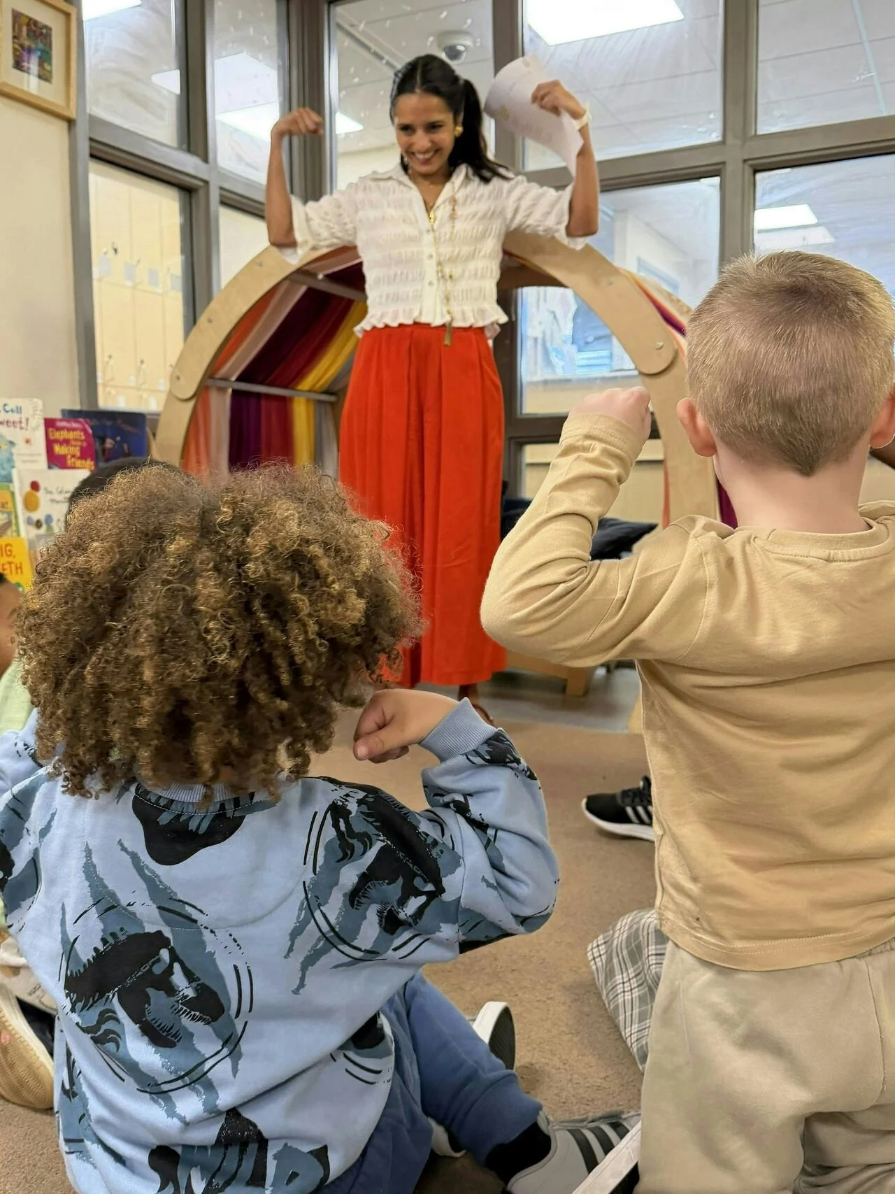 A woman dressed in a white top and bright orange skirt is standing on a raised platform, flexing her arms and smiling. She is holding a piece of paper in her right hand. Three children are sitting on the floor, watching her confidently. The setting appears to be a classroom or library with books and colorful background decorations.