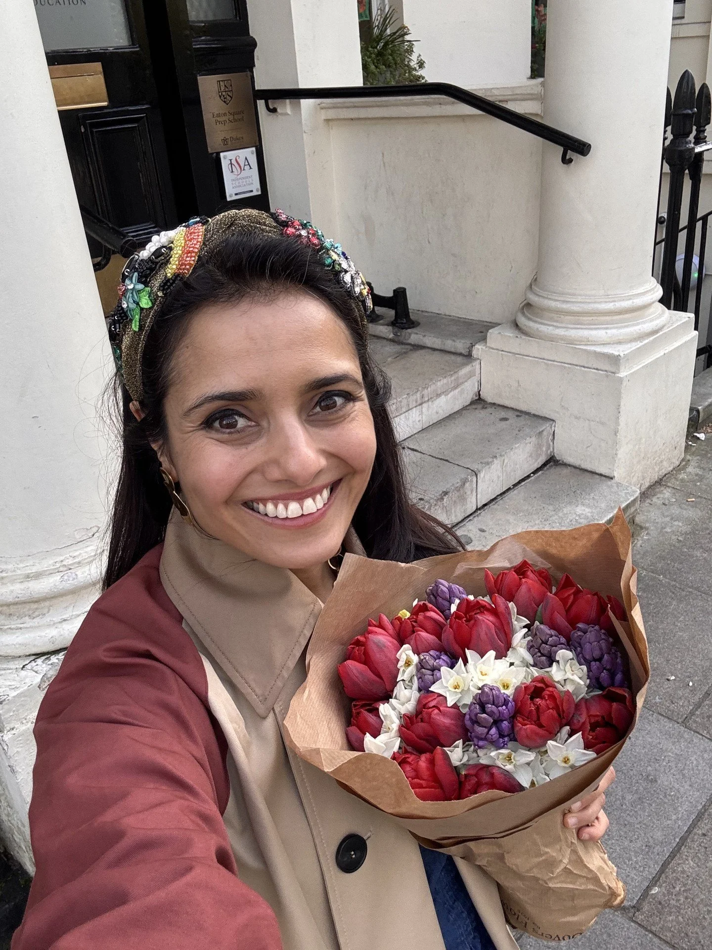 A woman is smiling and holding a bouquet of red, purple, and white flowers wrapped in brown paper, standing outside a white building with steps and a black railing.