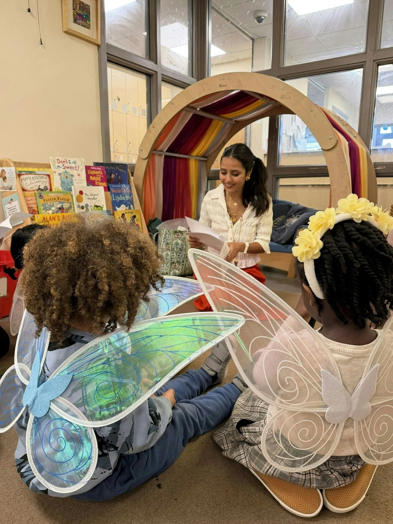 Young woman reading a book to children in costumes with fairy wings and antennae, inside a library or classroom setting.