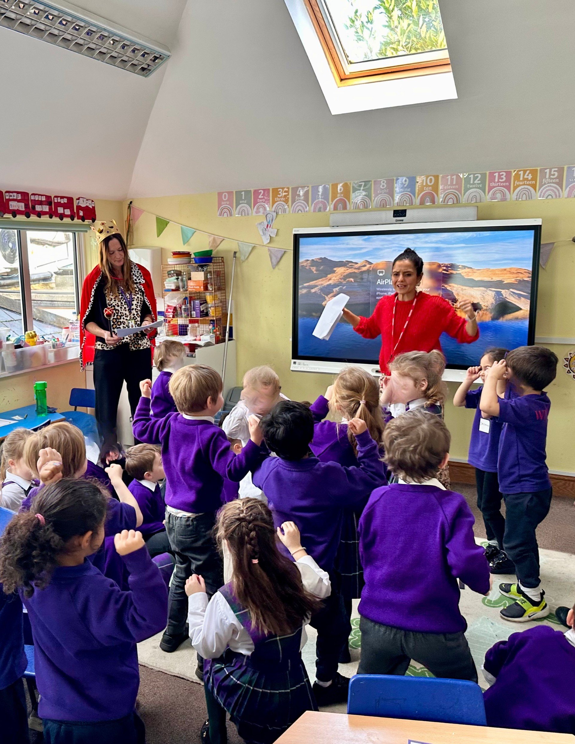 A classroom scene with children in purple uniforms gathered around and engaging with a teacher who is standing near a large screen display, with a background featuring a yellow wall decorated with numbered posters, hanging bunting, and a window with a view outside.