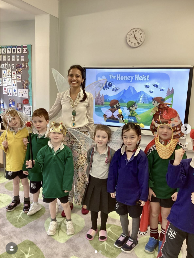 A teacher dressed as a fairy with fairy wings and a wand, standing with six children who are dressed in costumes for a play about bees and honey. The children are wearing crowns, animal-themed hats, and casual clothing. Behind them, a screen displays the title 'The Honey Heist,' and there is a classroom wall with educational posters.