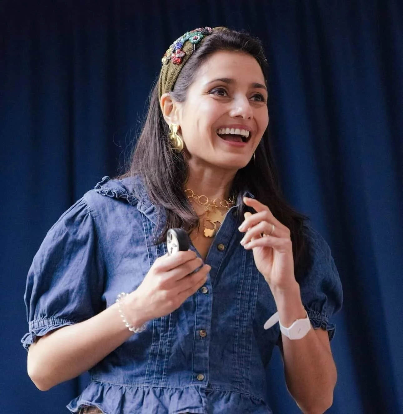 Woman with dark brown hair wearing a green headband with floral embellishments, gold earrings, necklace, wristwatch, and a blue denim dress, smiling and holding a microphone, standing in front of a dark curtain.