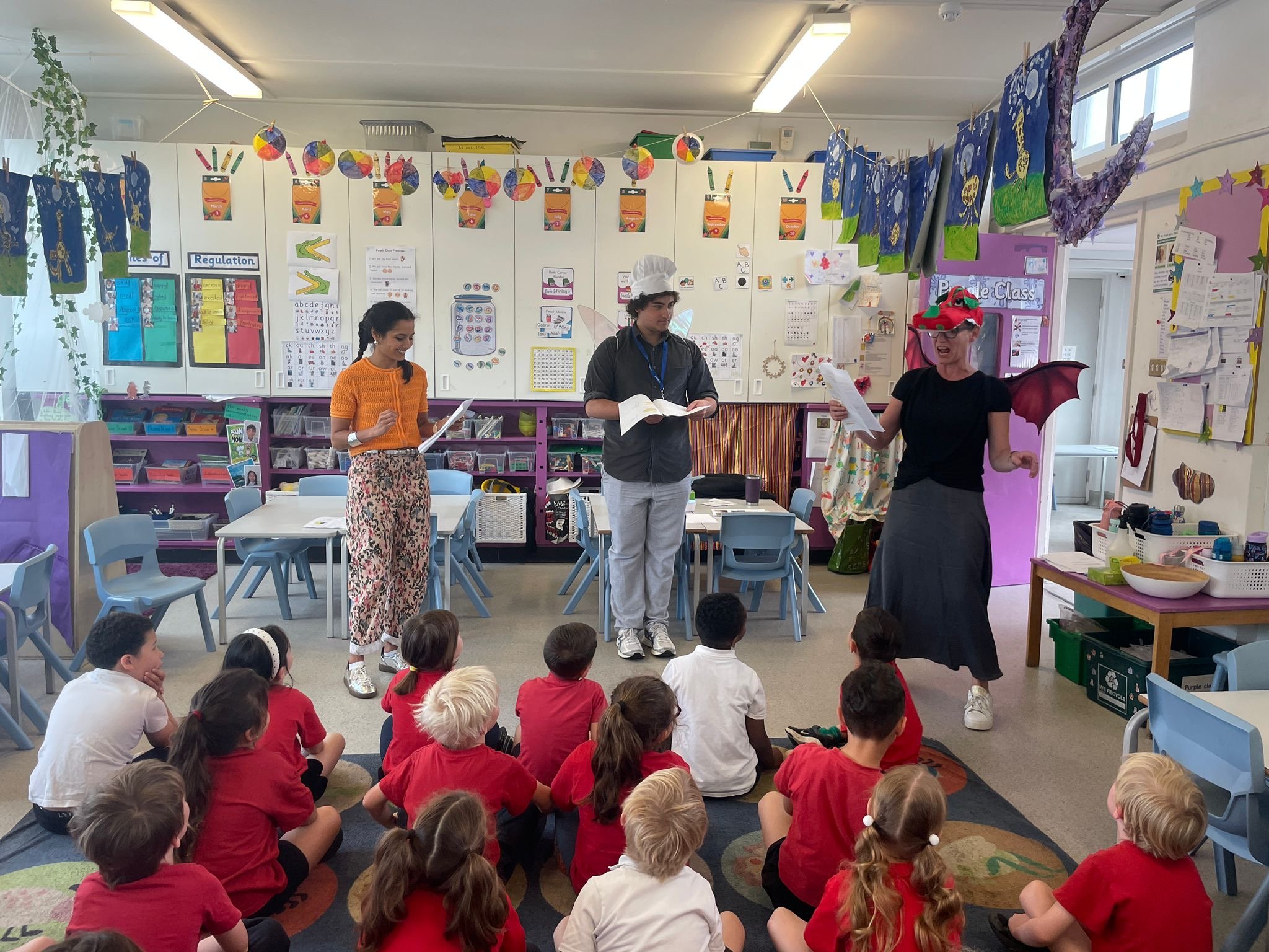 A classroom with children sitting on the floor and adults dressed in costumes standing at the front, likely performing for the children, with colorful decorations and educational posters on the walls.