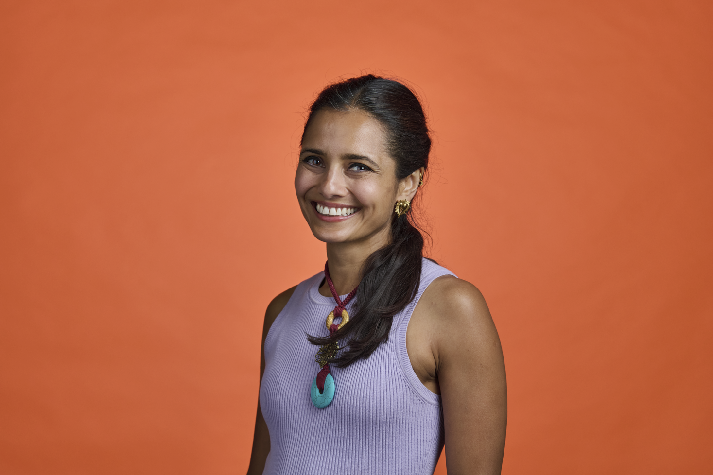 A smiling woman with dark hair tied back, wearing a light purple sleeveless top, gold earrings, and a colorful necklace, standing against an orange background.