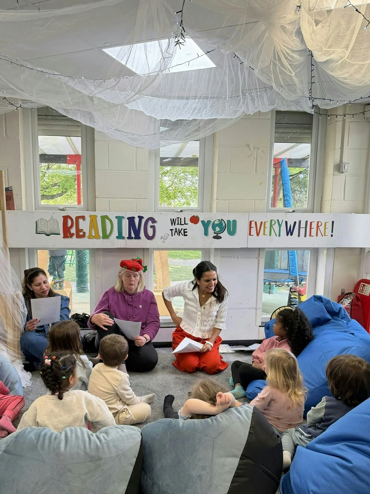 A group of children sitting on the floor listening to a woman reading in a library or classroom. There is colorful decor including a sign that says, 'READING WILL TAKE YOU EVERYWHERE!' over the window, which has white curtains and ceiling decorations. The woman leading the session is in a white top and orange pants, and children appear engaged and attentive.