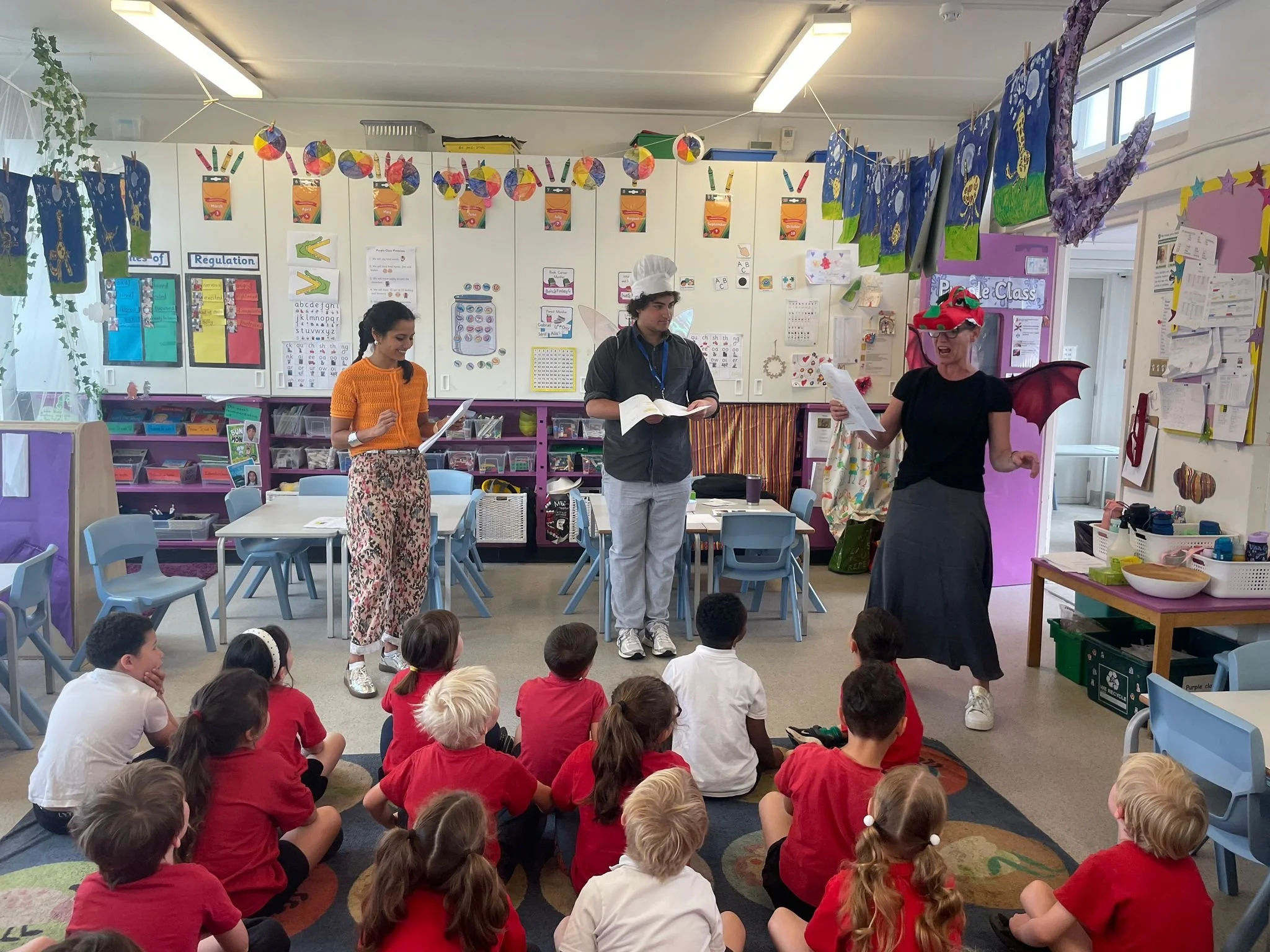 A classroom filled with young children sitting on the floor, listening to three people standing at the front, one dressed as a fairy, another as a chef, and the third as a dragon. The background has colorful posters, artwork, and educational decorations.
