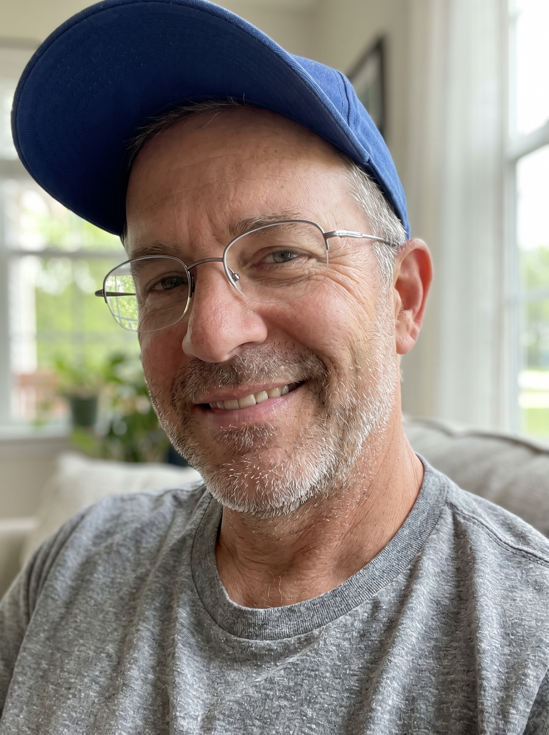Close-up of an older man smiling, with glasses, wearing a blue baseball cap and a gray t-shirt, inside a room near a window with curtains.