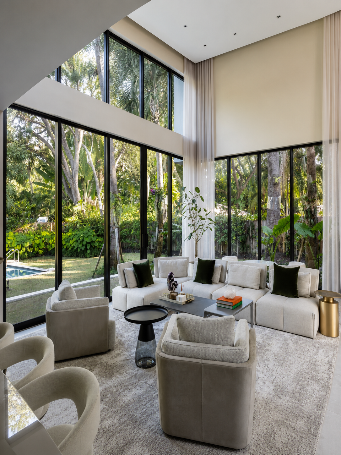 Modern living room with large floor-to-ceiling windows showing a lush green garden outside. The room has white cushioned sofas and armchairs, a black coffee table, and various decorative items including books, plants, and ornaments. Light curtains hang from the ceiling to the floor.