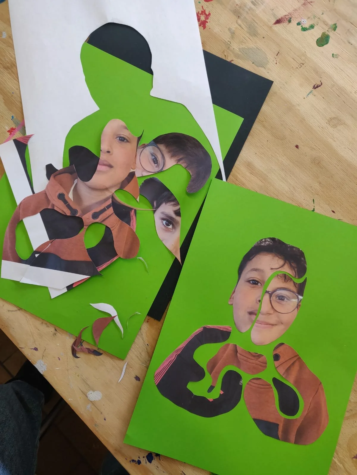 A collage art project with photos of children’s faces cut out and assembled into a dinosaur shape on a green background. Pieces of paper and photo fragments are scattered on a wooden table.