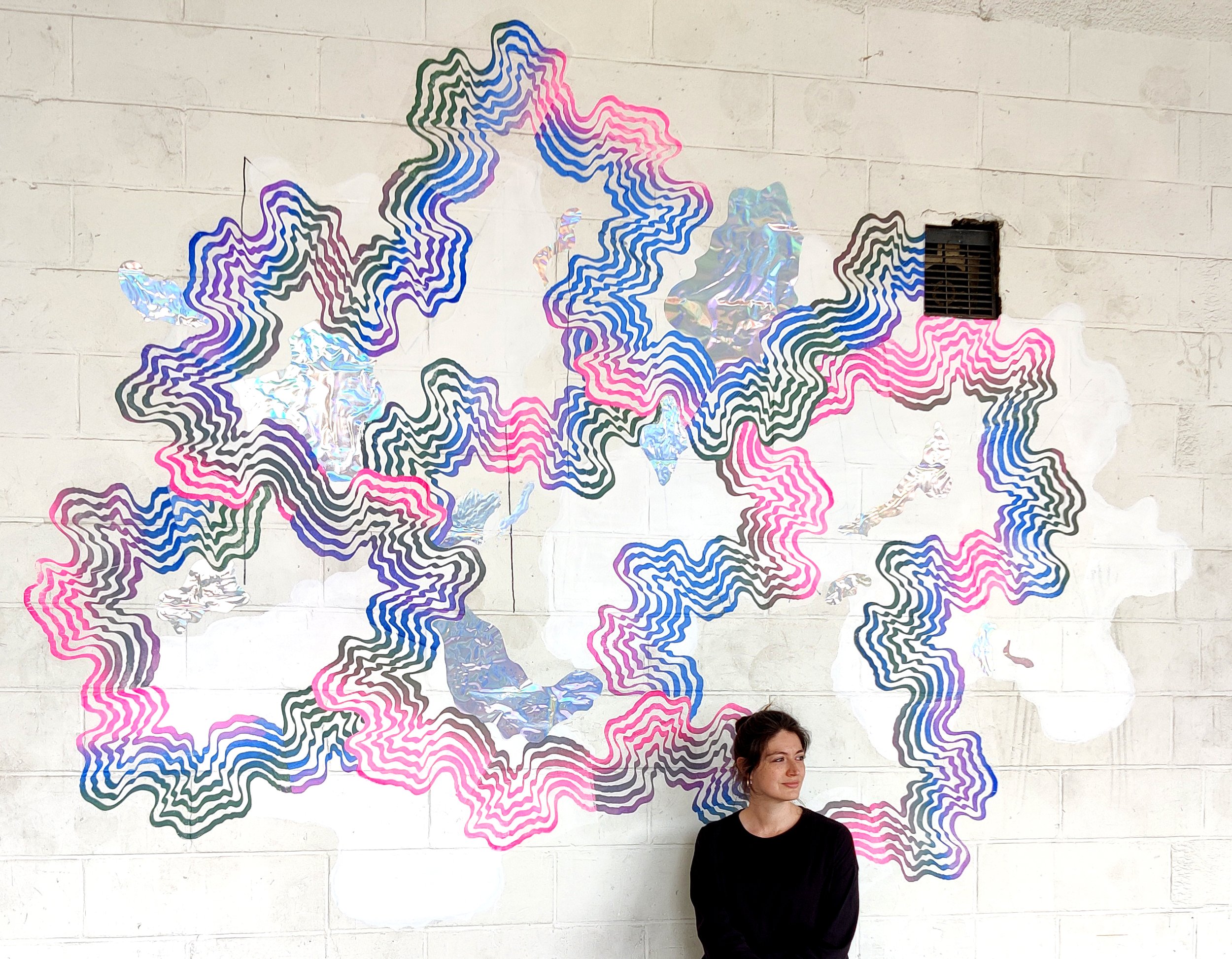A woman standing in front of a wall with colorful abstract line art that resembles a map of Germany, wearing a black top and looking to her left.