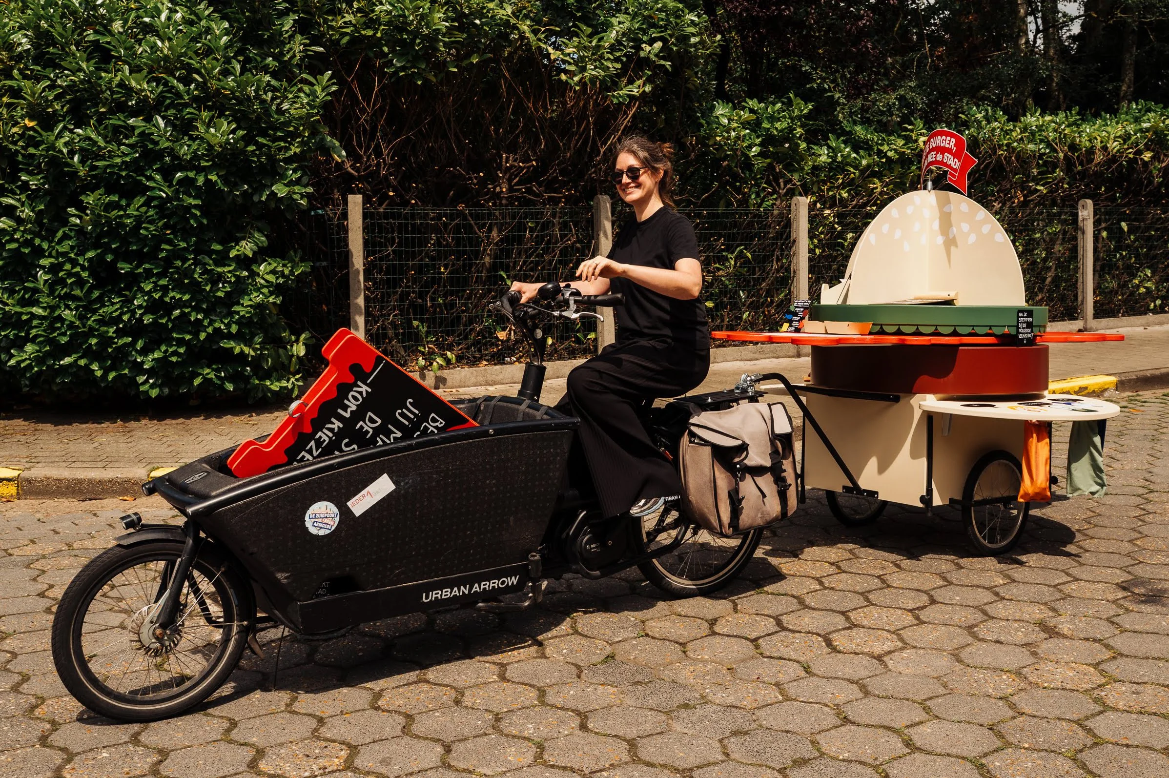 A woman riding a bicycle with a large frozen drink cart attached to the back. The woman is smiling, wearing sunglasses and a black shirt. The cart has a sign that says 'Yes, we deliver' and a big ice cream cone prop on top with a red sign that says 'Hamburger'. There is bushes and a fence in the background.