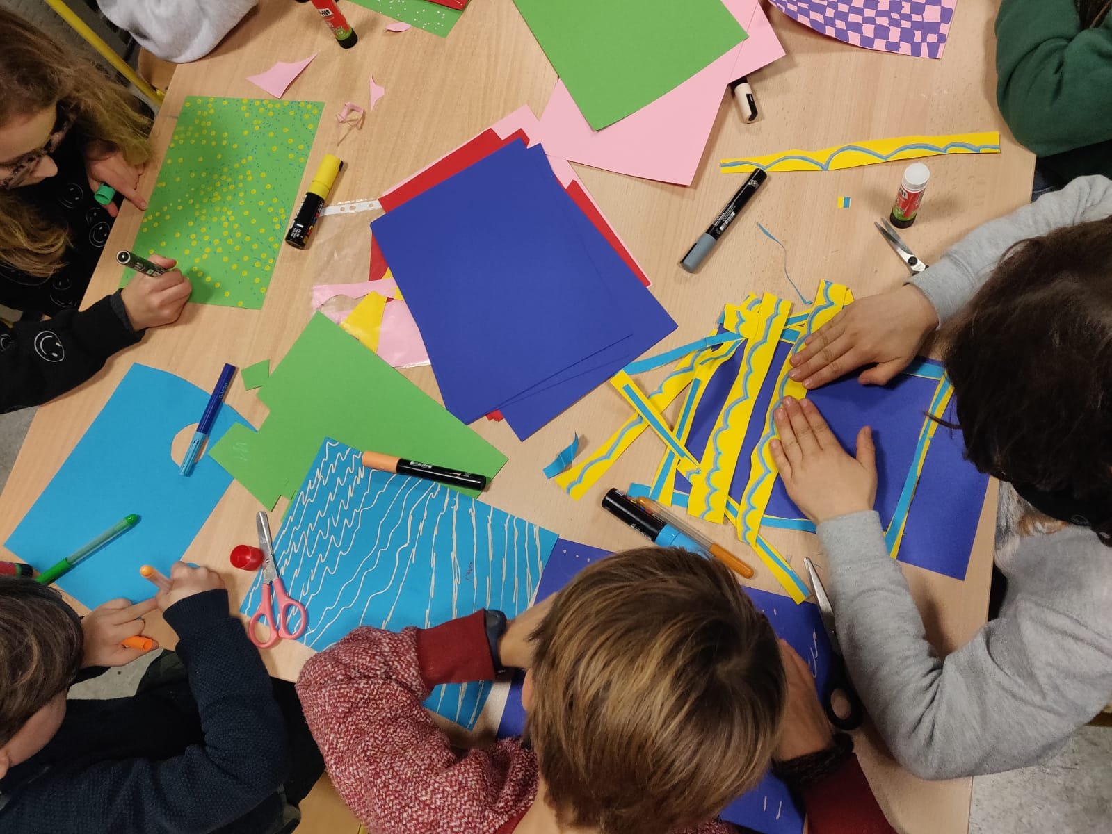 Children creating arts and crafts with colorful paper, scissors, markers, tape, and glue on a wooden table.