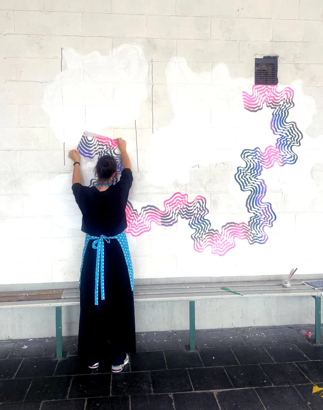 A person with short hair, wearing a black top and a long black skirt with a blue polka dot apron, is drawing on a large white wall with a black marker. The wall features an abstract art piece with wavy, concentric lines in pink, gray, and blue, organ