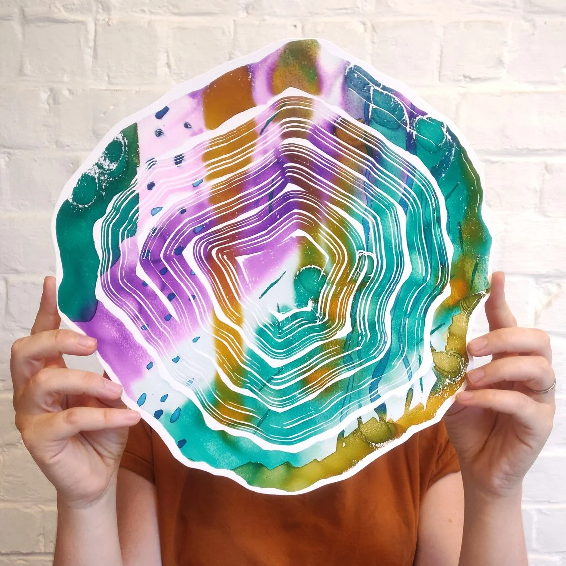 Person holding a colorful abstract piece of art resembling a topographical map or geological formation, with concentric white lines and vibrant hues of purple, green, yellow, and brown, against a white brick wall background.
