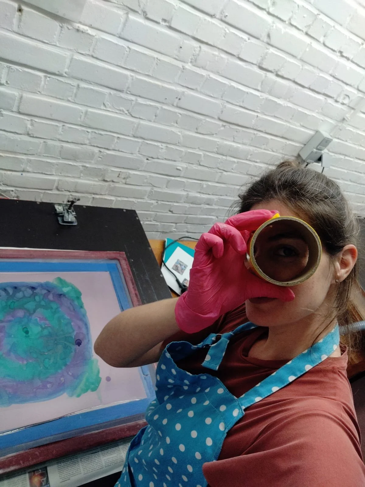 A young woman wearing a blue polka dot apron, pink gloves, and a brown shirt looking through a magnifying glass at water-colored artwork of a jellyfish on paper, with a white painted brick wall in the background.
