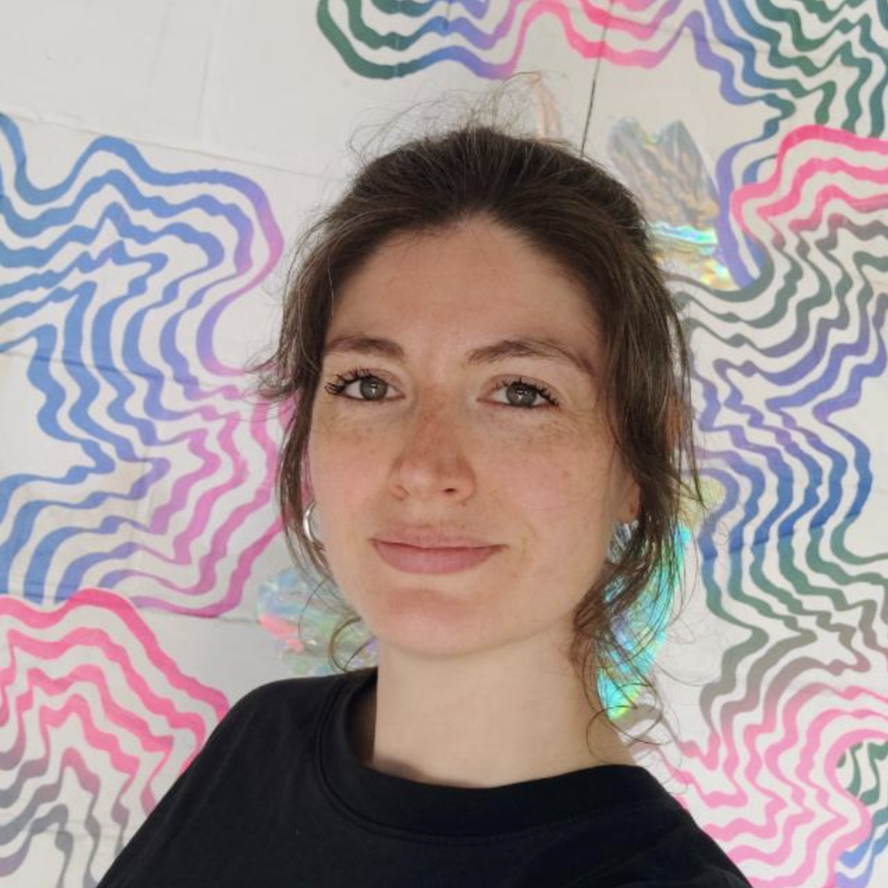 A young woman with brown hair, freckles, and earrings, smiling in front of a colorful, abstract, wavy pattern background.