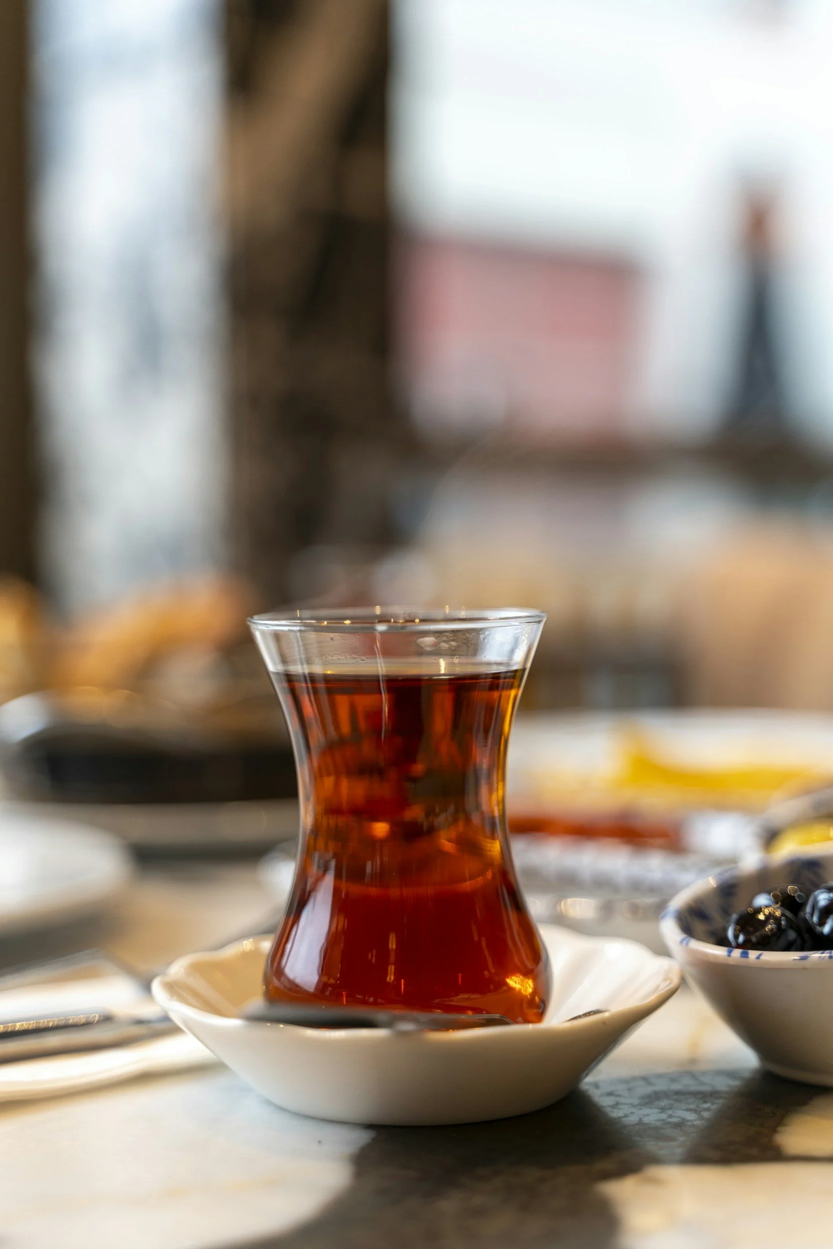 Cam bir fincanda Türk çayı, altında küçük bir beyaz tabak var ve yanında küçük kaselerde meyve veya kuru yemişler bulunuyor.