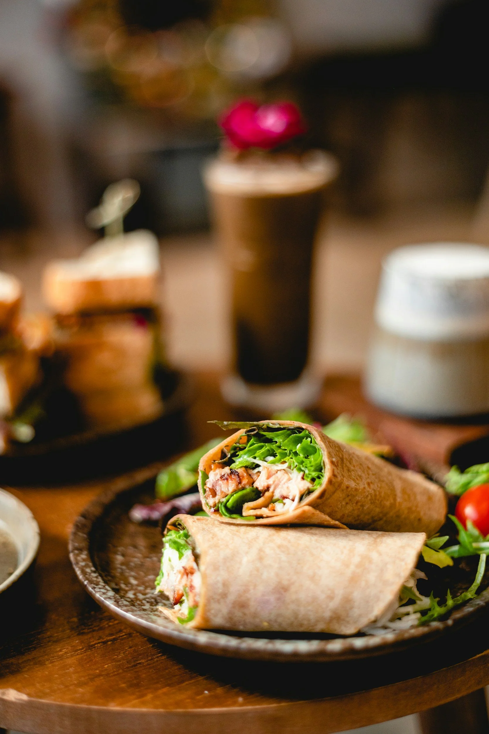 Bir tabakta dürüm burrito, içinde marul, tavuk ve diğer malzemeler var. Arka planda barda ve vazo içinde çiçekler bulunmaktadır.