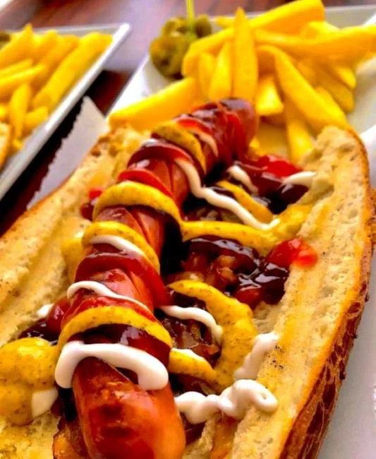 Büyük boy hot dog ekmek içinde sosis, üzerinde ketçap, hardal, mayonez ve turşu dilimleri ile hazırlanmış. Yanında patates kızartması bulunan tabakta.