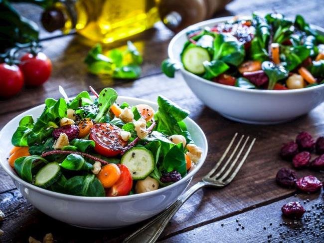 İki adet büyük kase içinde taze sebze salatası, bir tanesinde salata dolu, diğerinde ise sadece sebzeler. Masada çatal ve birkaç kuru kayısı var. Arka planda zeytinyağı şişesi ve domatesler.